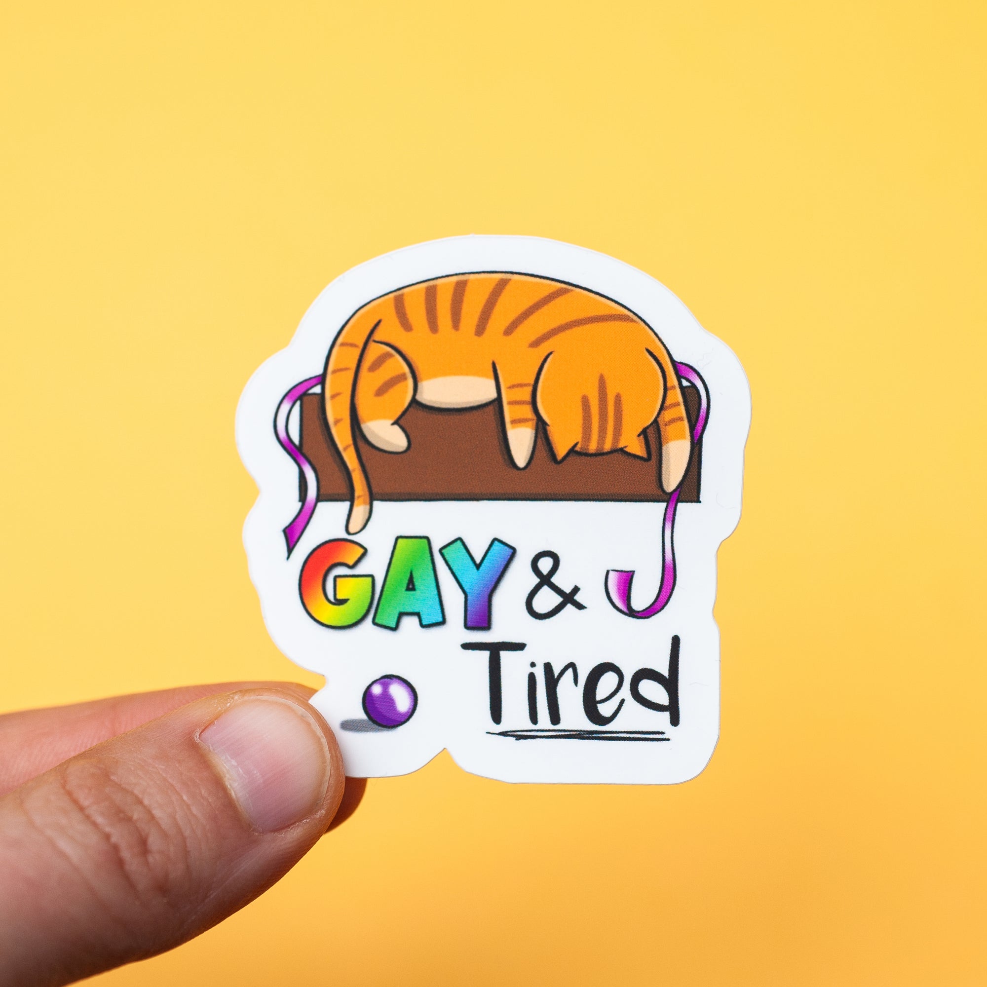 An orange tabby cat folded over a table, looking asleep, with a ball down on the floor & the text "gay & tired"